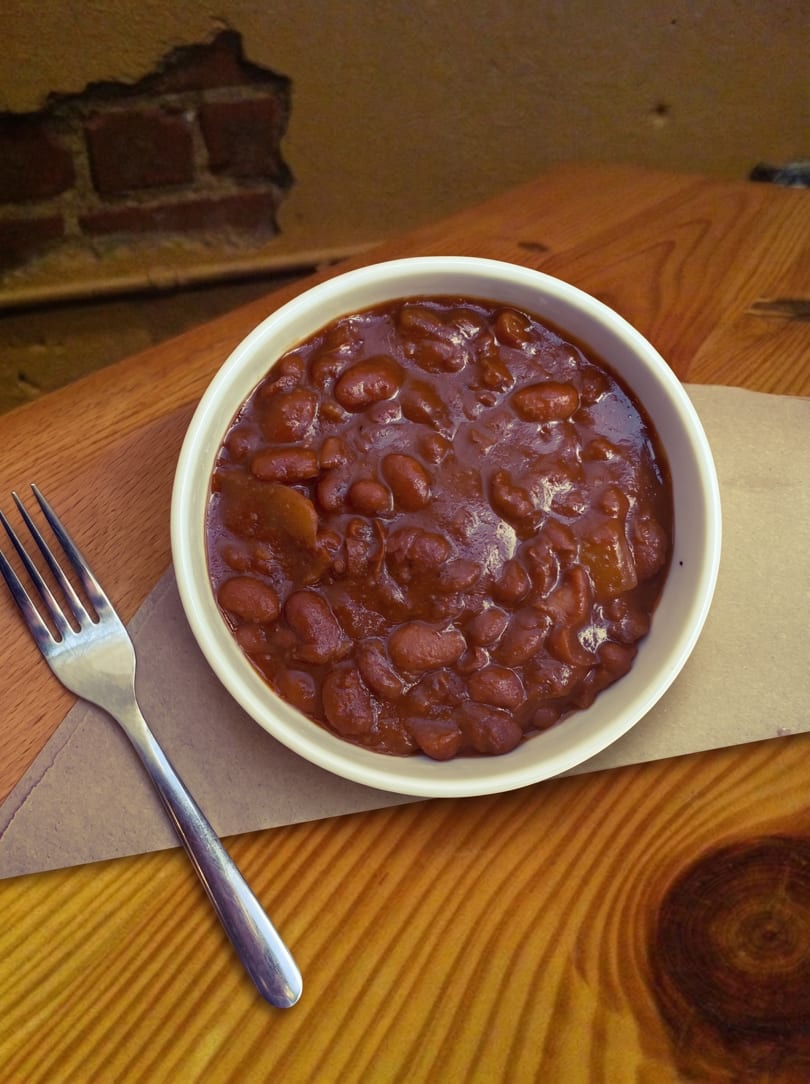 A white bowl of baked beans with a fork on a brown paper napkin and wooden table.