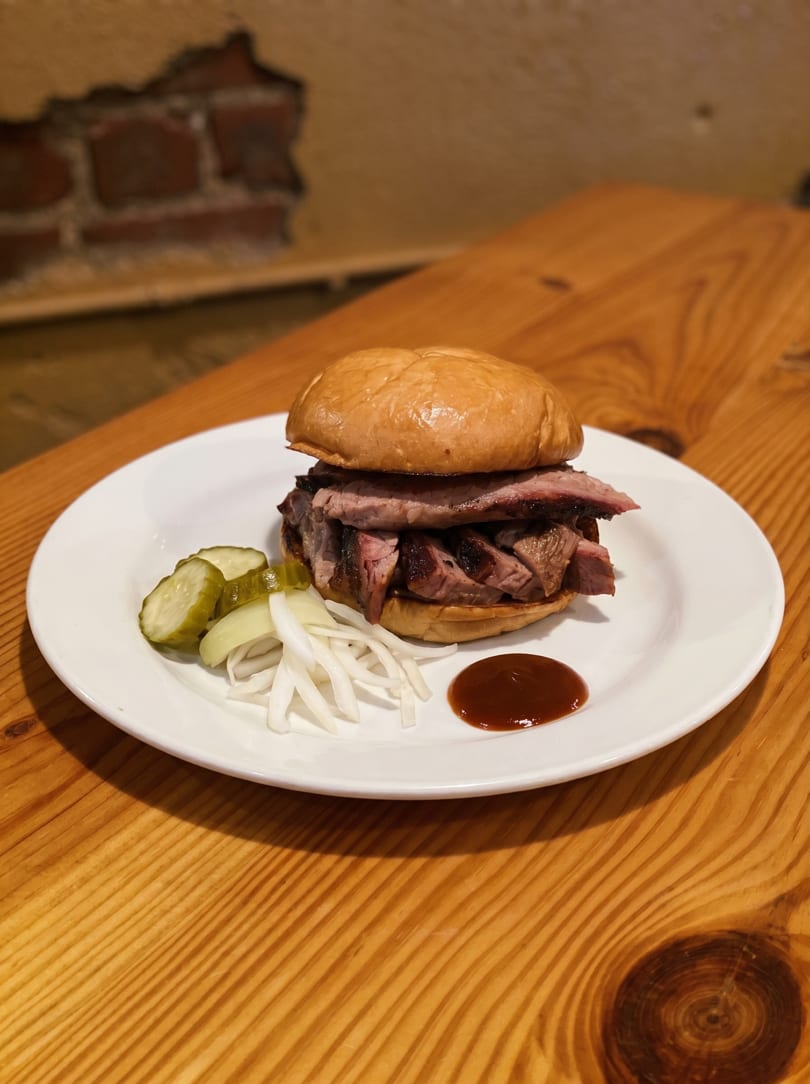 A brisket sandwich with pickles, onions, and barbecue sauce on a white plate on a wooden table.