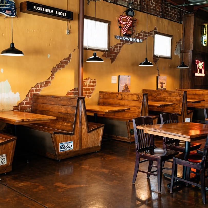 Restaurant interior with wooden booths and tables, exposed brick, and neon signs on a distressed yellow wall.