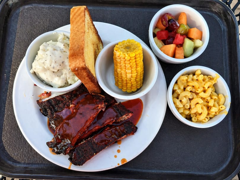 BBQ ribs with mashed potatoes, corn on the cob, mac and cheese, and fruit salad.
