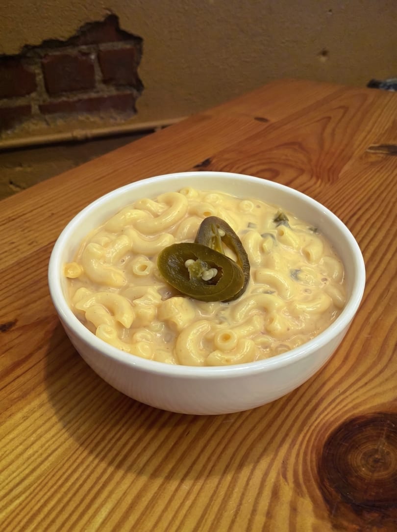 Creamy macaroni and cheese in a white bowl, topped with two sliced jalapeños, on a wooden table.
