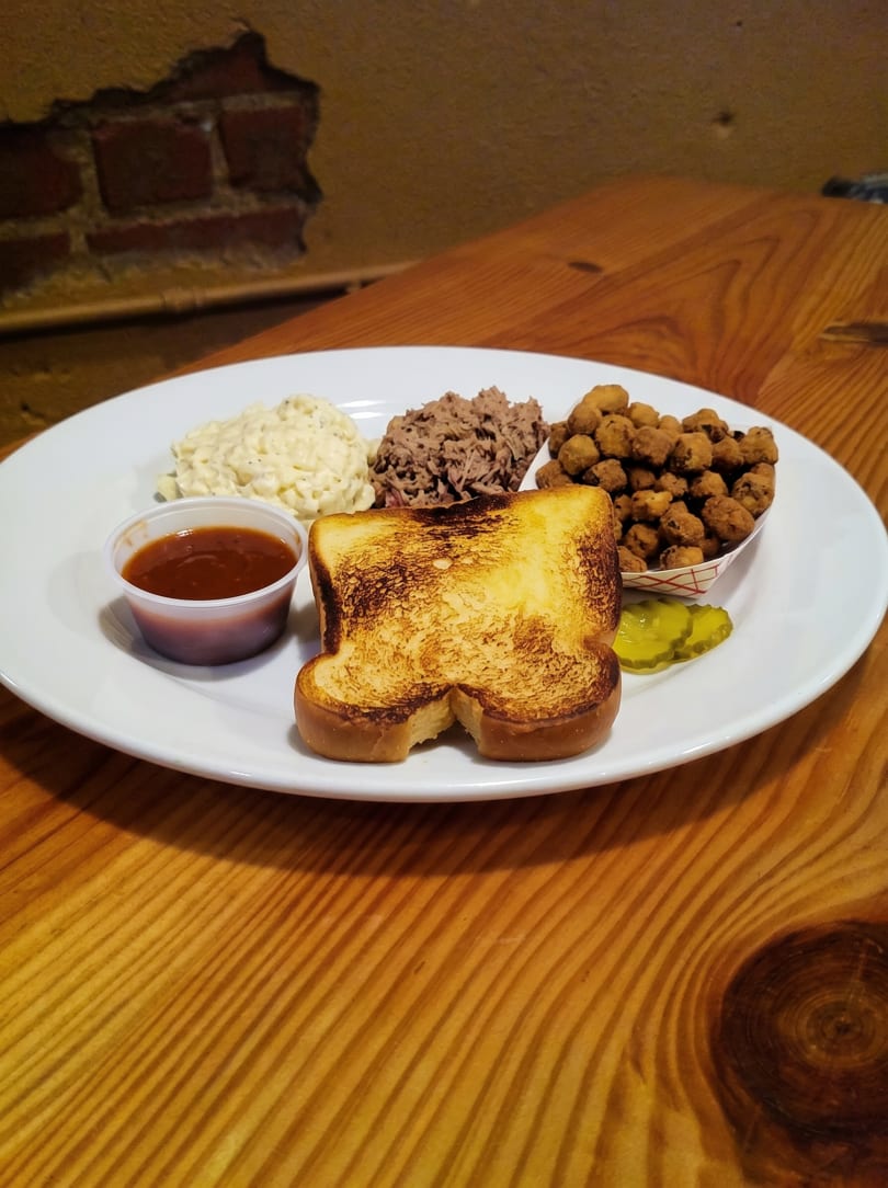 A BBQ plate with pulled pork, potato salad, fried okra, toasted bread, pickles, and sauce.