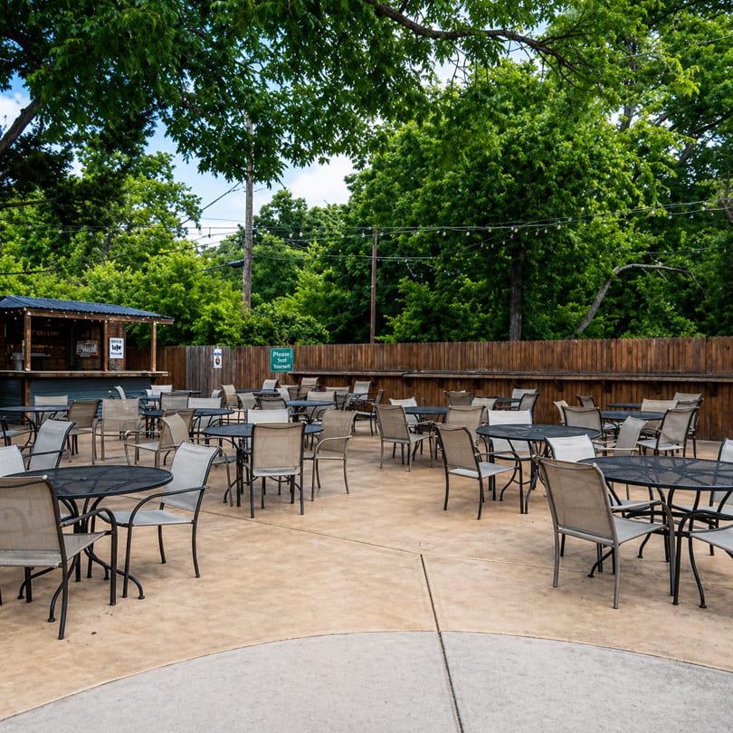 Outdoor patio with numerous tables and chairs, a bar, and lush green trees under string lights.