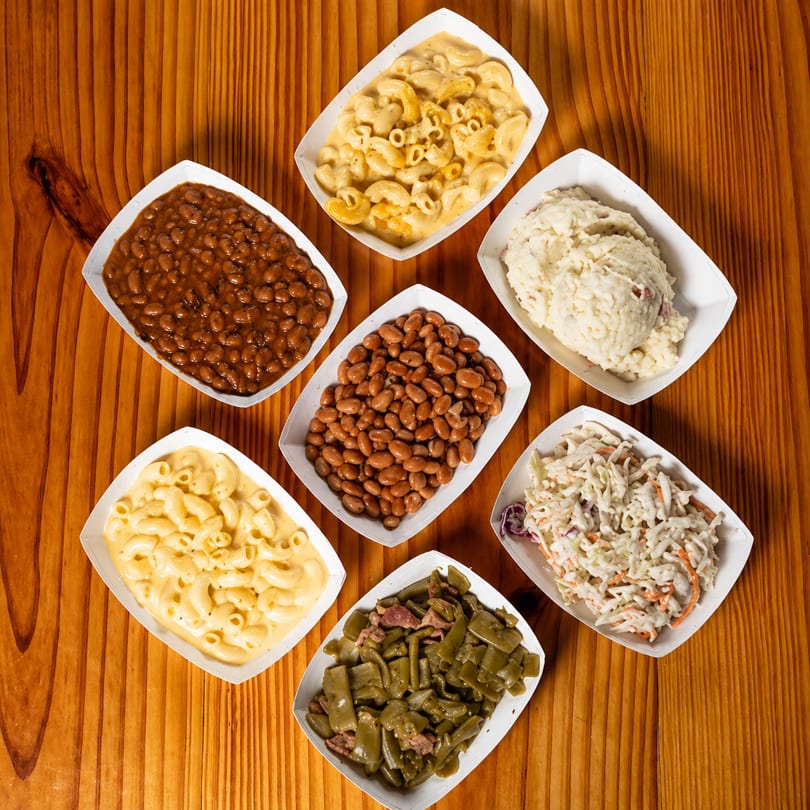 Seven side dishes, including mac and cheese, baked beans, and coleslaw, on a wooden table.